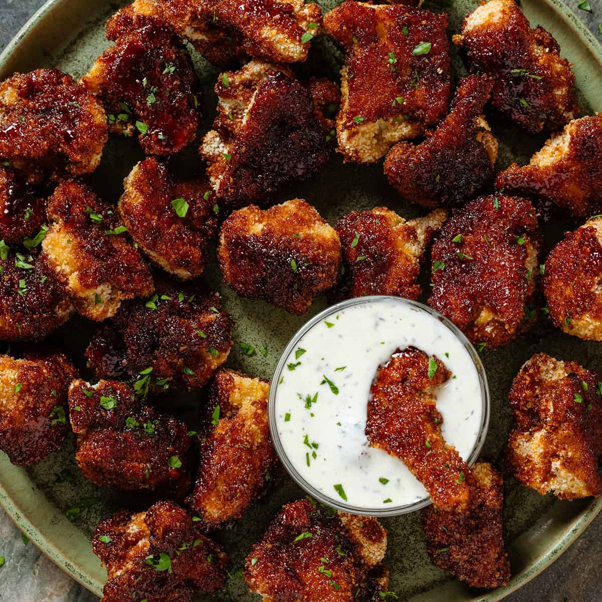 Baked breadcrumbed bbq cauliflower florets on a green plate with a small glass bowl of white dip in the middle with one floret dipped in it.