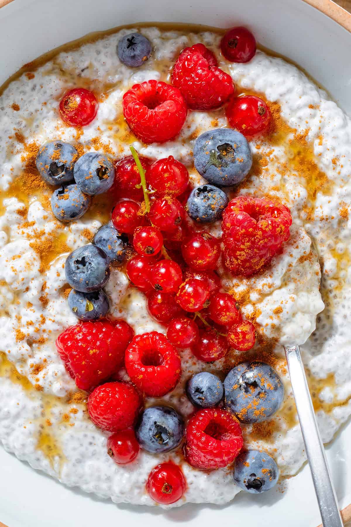 White Greek yogurt chia pudding topped with fresh blueberries, raspberries, currants, maple syrup, and cinnamon in a white bowl with a silver spoon on the right in the bowl.