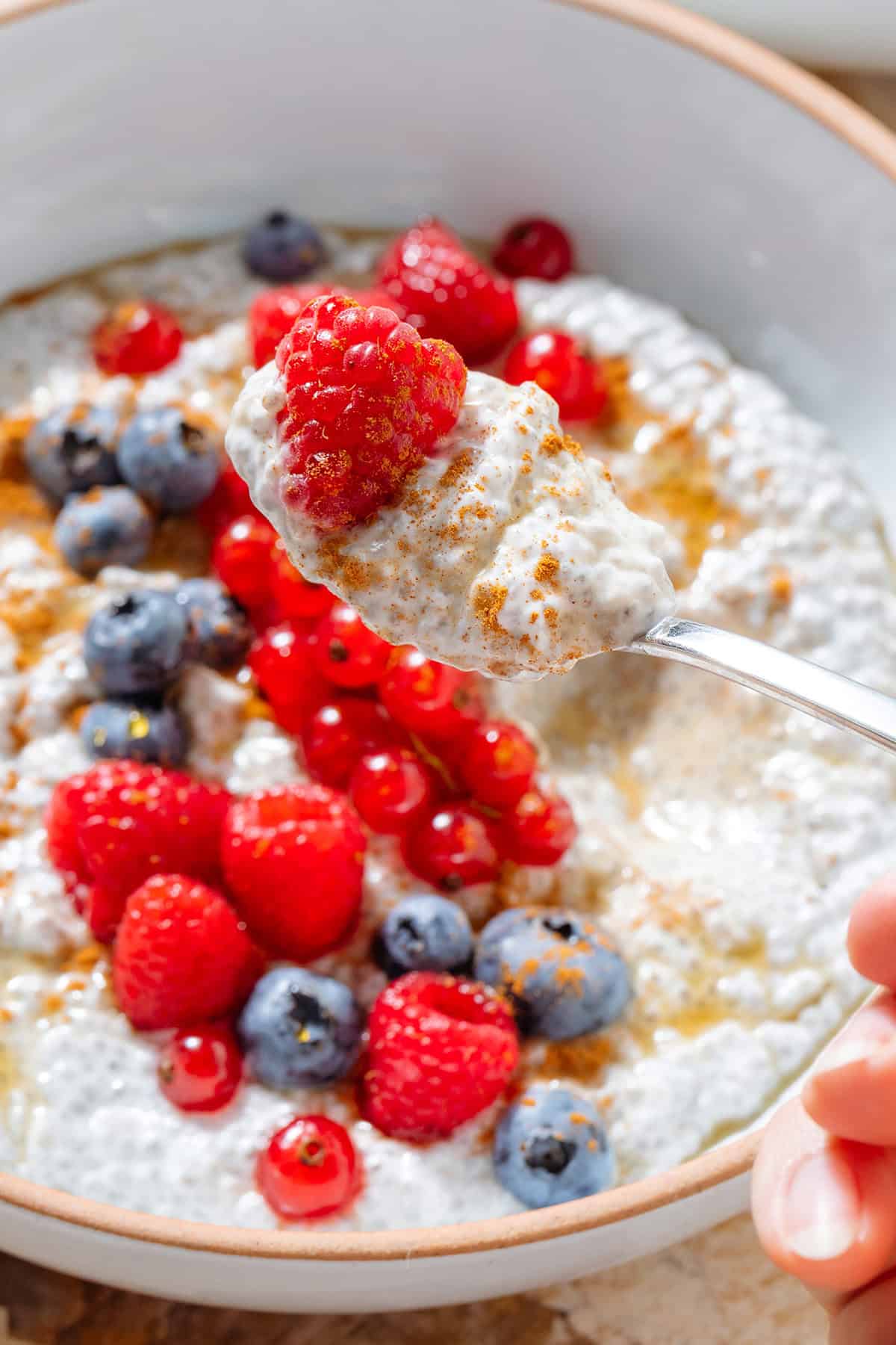 A hand holding a spoon with chia pudding and a raspberry over a white bowl with white chia pudding topped with berries.