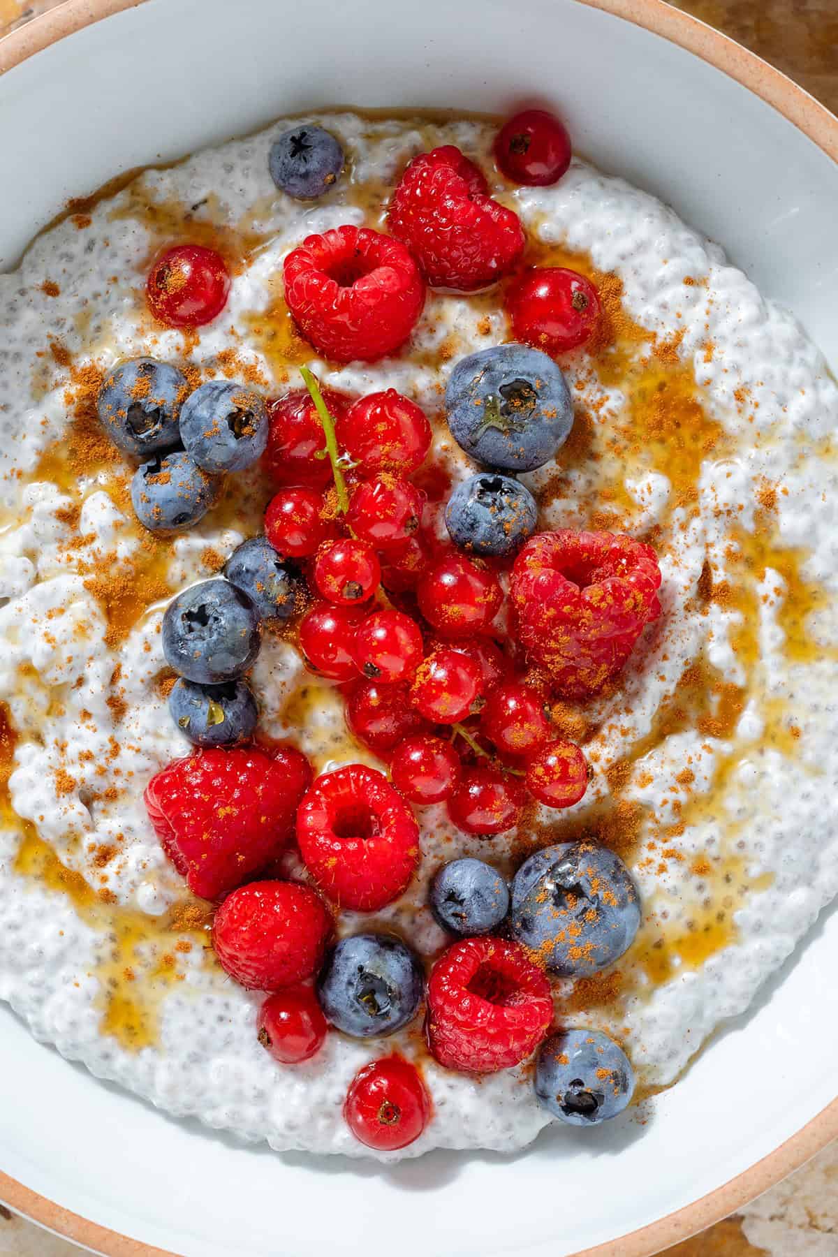 White Greek yogurt chia pudding topped with fresh blueberries, raspberries, currants, maple syrup, and cinnamon in a white bowl with a beige rim.