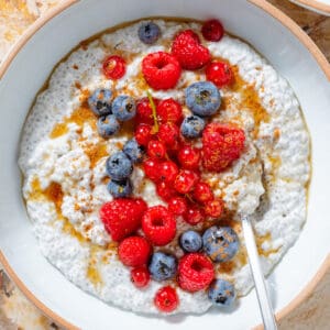 White Greek yogurt chia pudding topped with fresh blueberries, raspberries, currants, maple syrup, and cinnamon in a white bowl with a silver spoon.