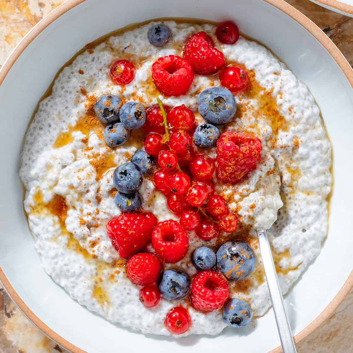 White Greek yogurt chia pudding topped with fresh blueberries, raspberries, currants, maple syrup, and cinnamon in a white bowl with a silver spoon.