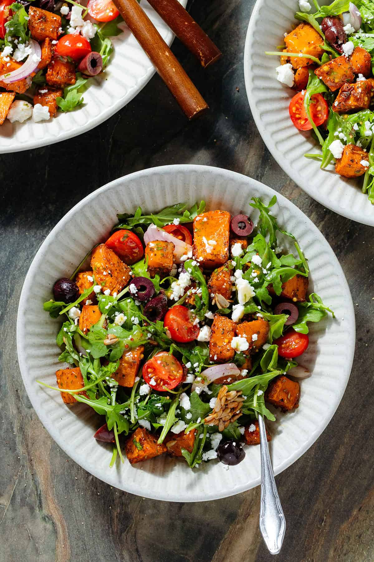 Three white bowls with arugula and sweet potato salad topped with feta, cherry, tomatoes, olives, and roasted sunflower seeds with a fork in the bowl on the bottom.