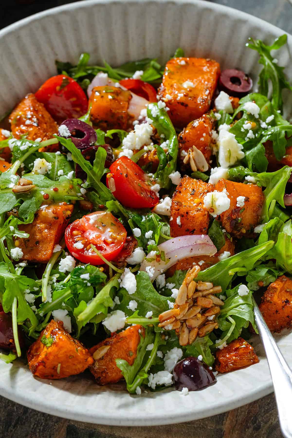 Arugula salad with diced roasted sweet potatoes with kalamata olives, feta, cherry tomatoes, sunflower seeds, and red onion drizzled with vinaigrette in a white bowl.
