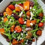 Arugula salad with diced roasted sweet potatoes with kalamata olives, feta, cherry tomatoes, sunflower seeds, and red onion drizzled with vinaigrette in a white bowl.