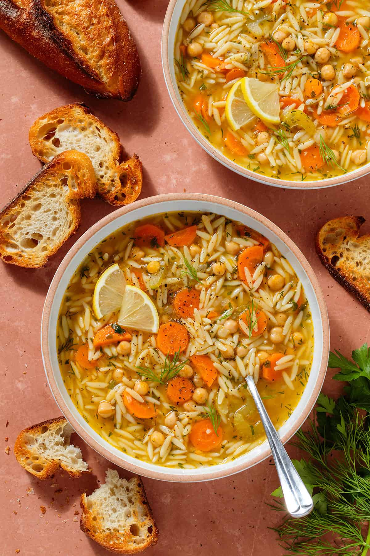 Orzo soup with carrots and chickpeas in a low bowl garnished with lemon triangles and with a spoon.