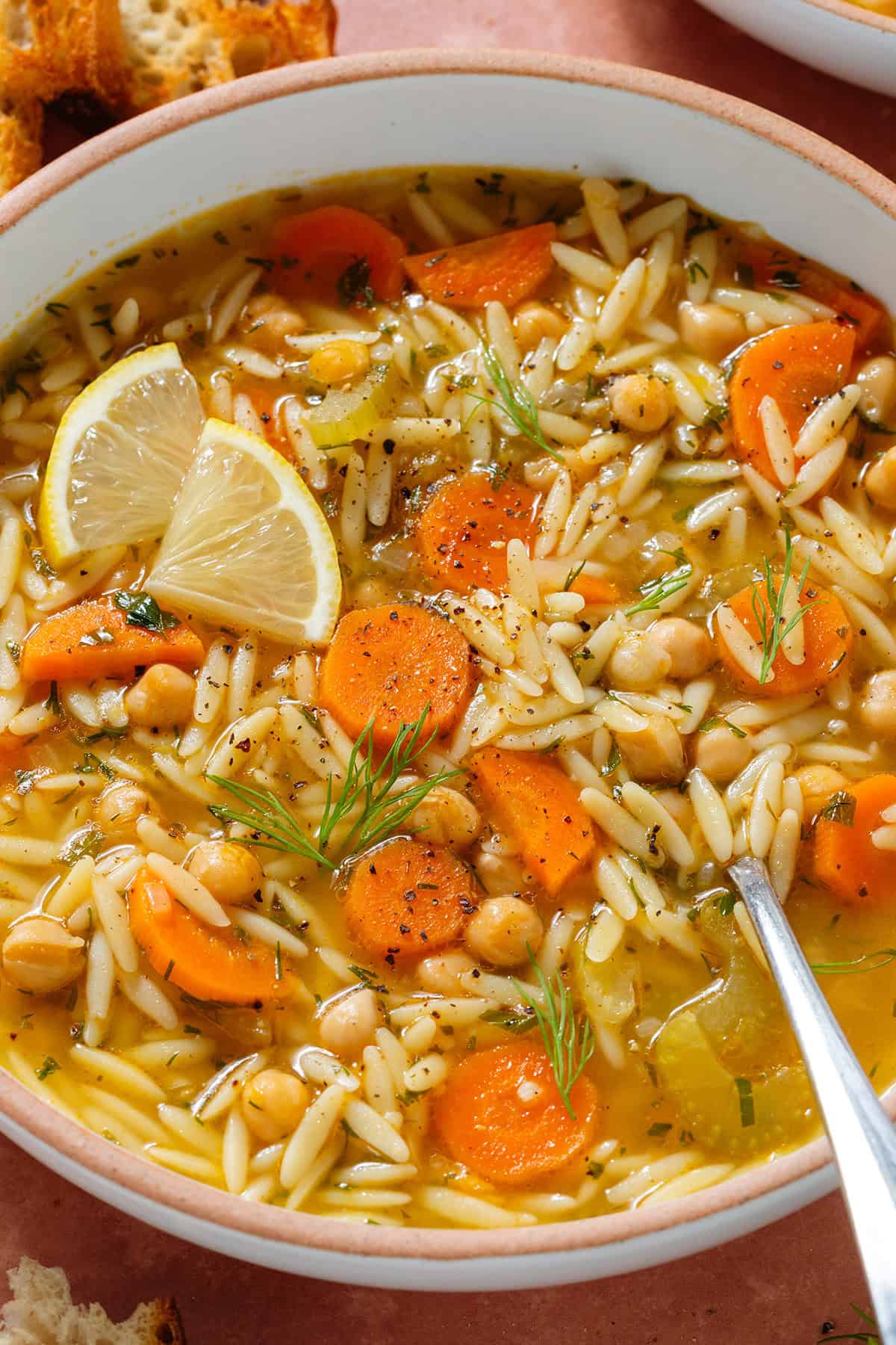 Orzo soup with carrots and chickpeas in a low bowl garnished with lemon triangles, black pepper, and fresh dill.