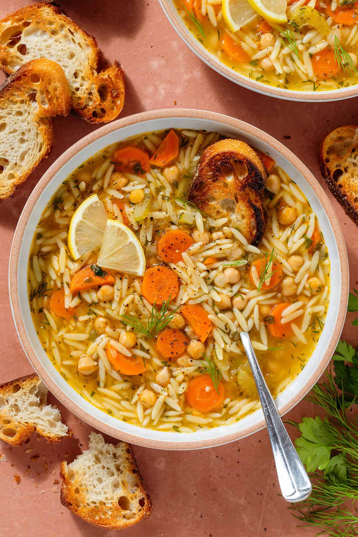 Orzo soup with carrots and chickpeas in a low bowl garnished with lemon triangles a spoon and a slice of toasted baguette dipped in.