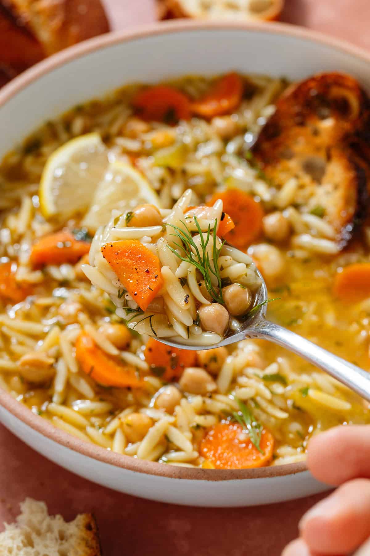 A spoon scooping orzo soup with carrots and chickpeas in a low bowl garnished with lemon triangles, black pepper, and fresh dill.