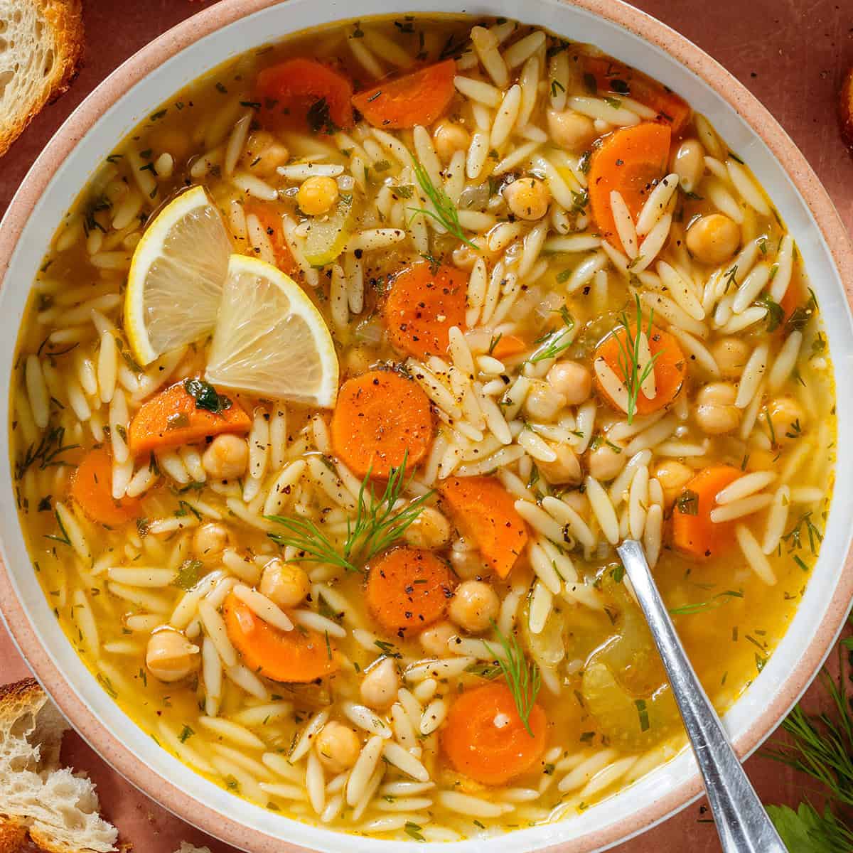 Orzo soup with carrots and chickpeas in a low bowl garnished with lemon triangles, black pepper, and fresh dill.
