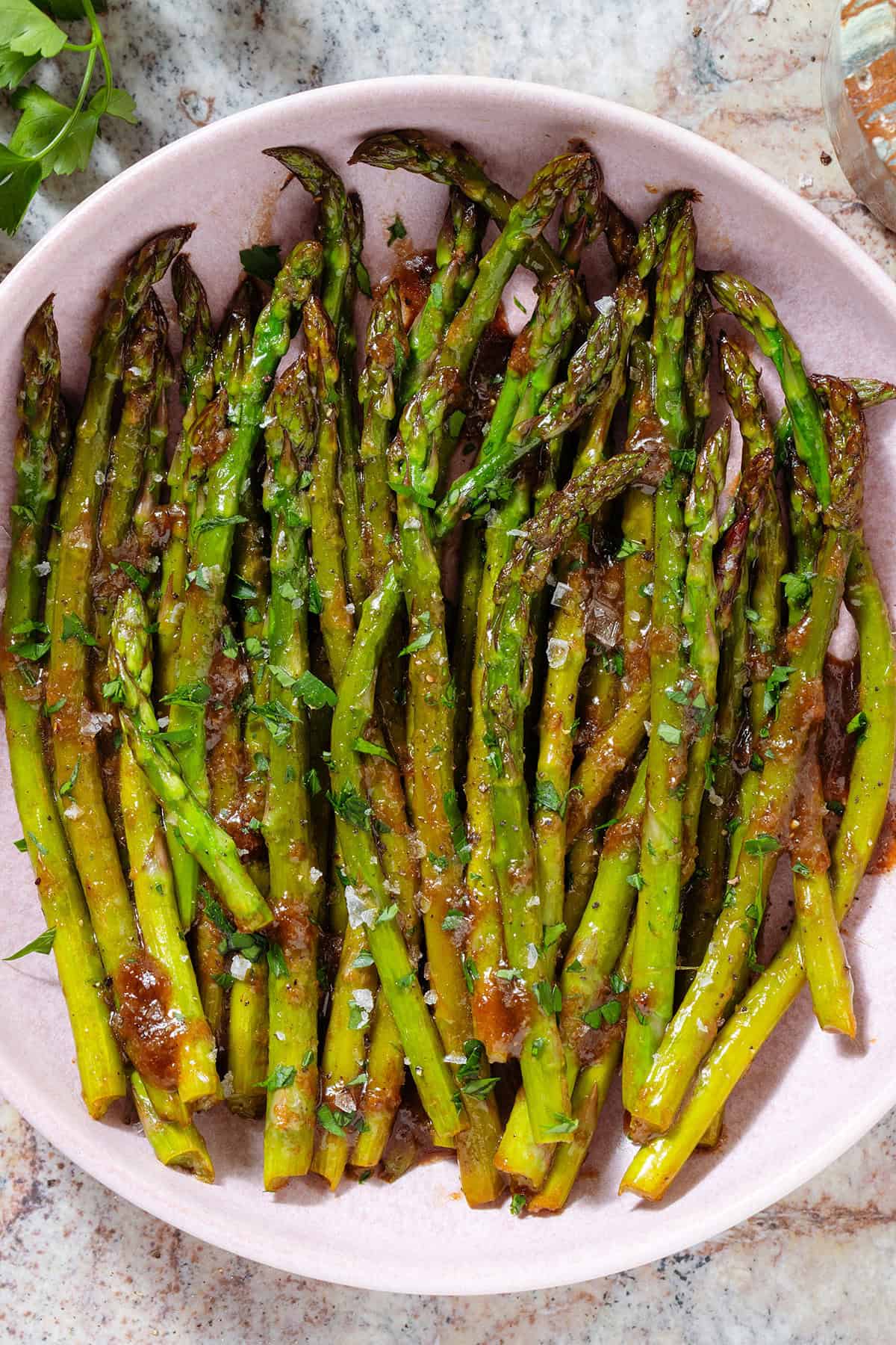 Sauteed balsamic glazed asparagus in a pink low bowl sprinkled with flaky sea salt, black pepper, and chopped fresh parsley.