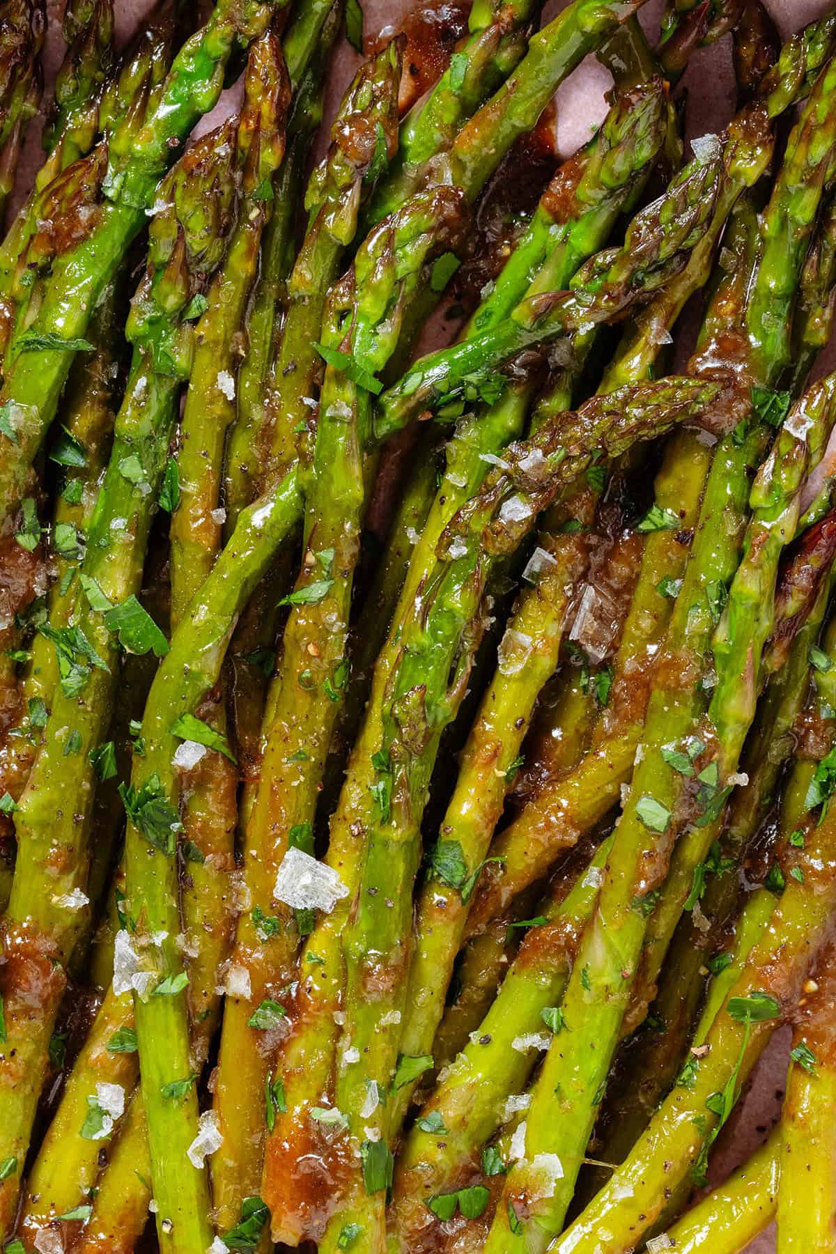 Sauteed balsamic glazed asparagus in a pink low bowl sprinkled with flaky sea salt, black pepper, and chopped fresh parsley.