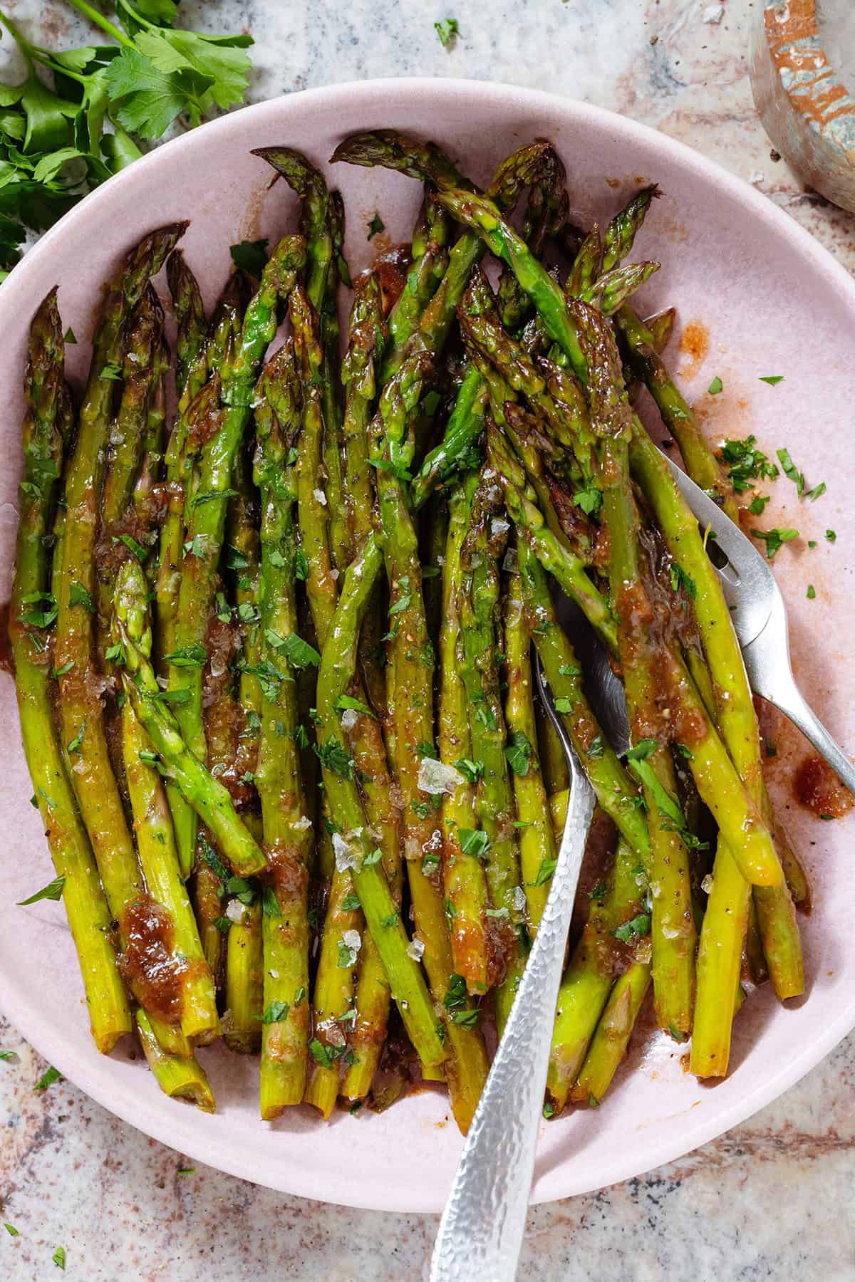 Sauteed balsamic glazed asparagus in a pink low bowl sprinkled with flaky sea salt, black pepper, and chopped fresh parsley with two forks in the bowl.