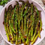 Sauteed balsamic glazed asparagus in a pink low bowl sprinkled with flaky sea salt, black pepper, and chopped fresh parsley.