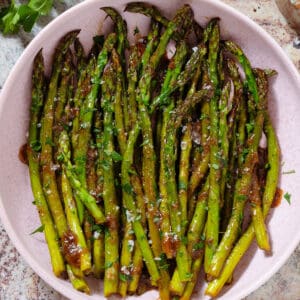 Sauteed balsamic glazed asparagus in a pink low bowl sprinkled with flaky sea salt, black pepper, and chopped fresh parsley.