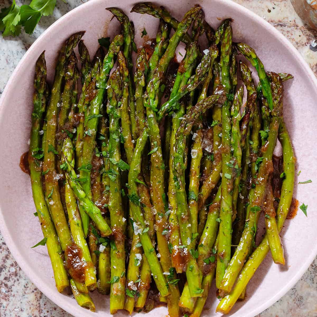 Sauteed balsamic glazed asparagus in a pink low bowl sprinkled with flaky sea salt, black pepper, and chopped fresh parsley.