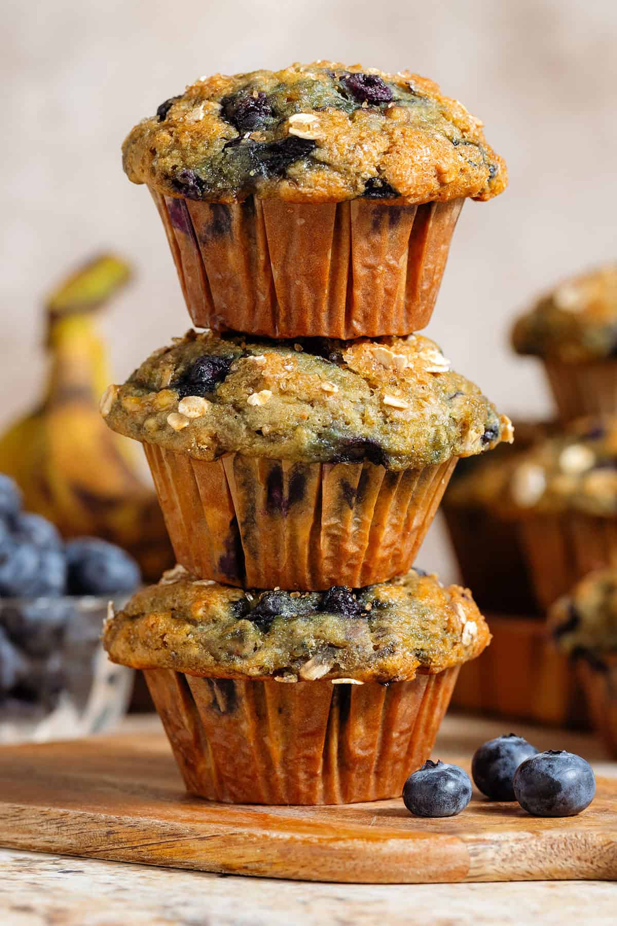 Three blueberry banana muffins with oats stacked on a small wooden cutting board.