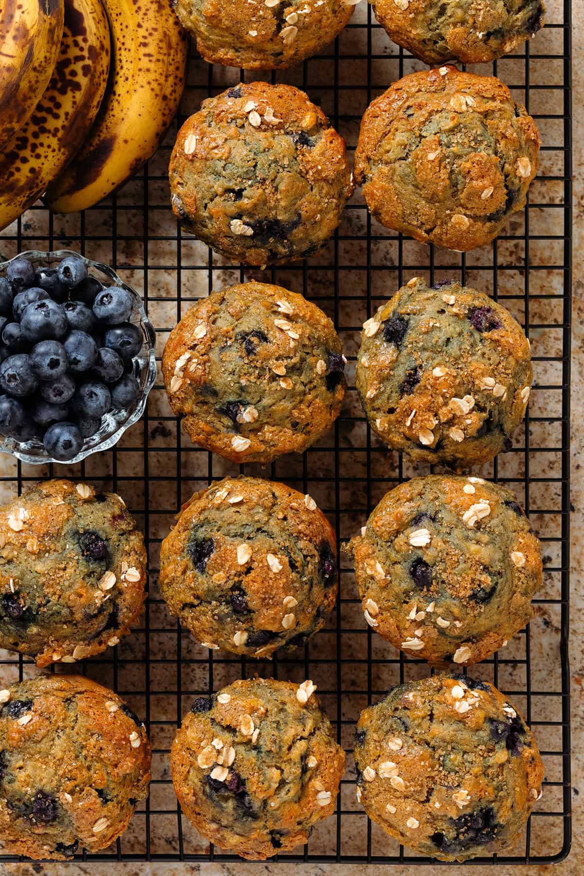 Banana blueberry oat muffins on a black wire rack with fresh blueberries in a small glass bowl and ripe bananas in the corner.