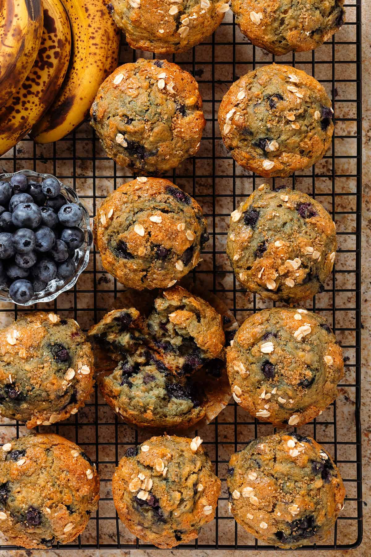 Banana blueberry oat muffins and fresh blueberries on a black wire rack with the muffin in the middle sliced in half showing the soft inside.