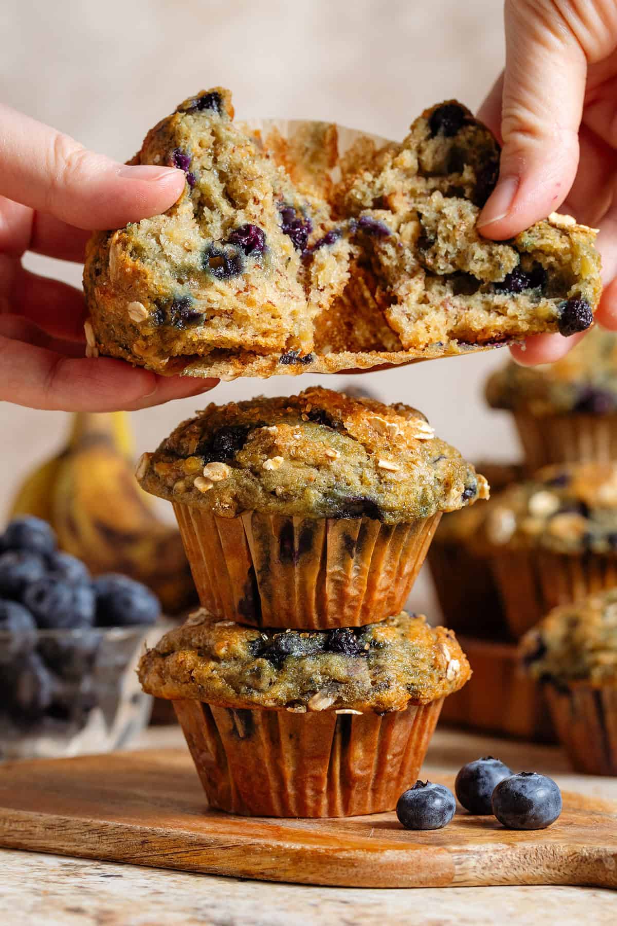 Two blueberry oatmeal muffins stacked and two hands holding one muffin torn open over them to show the fluffy inside.