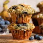 Two blueberry banana muffins with oats stacked on a small wooden cutting board.