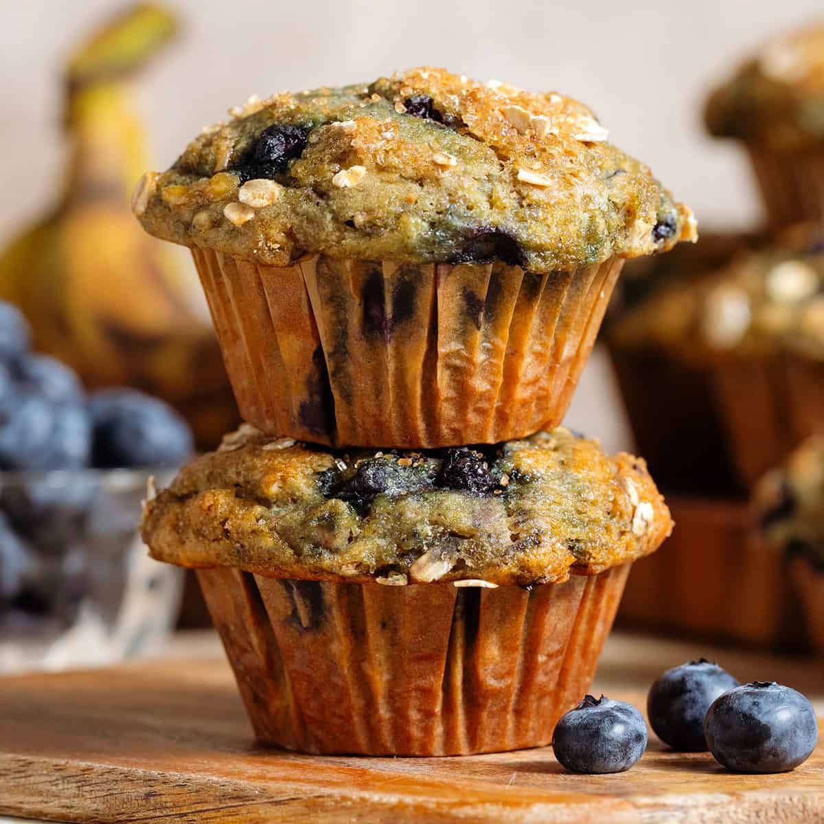 Two blueberry banana muffins with oats stacked on a small wooden cutting board.
