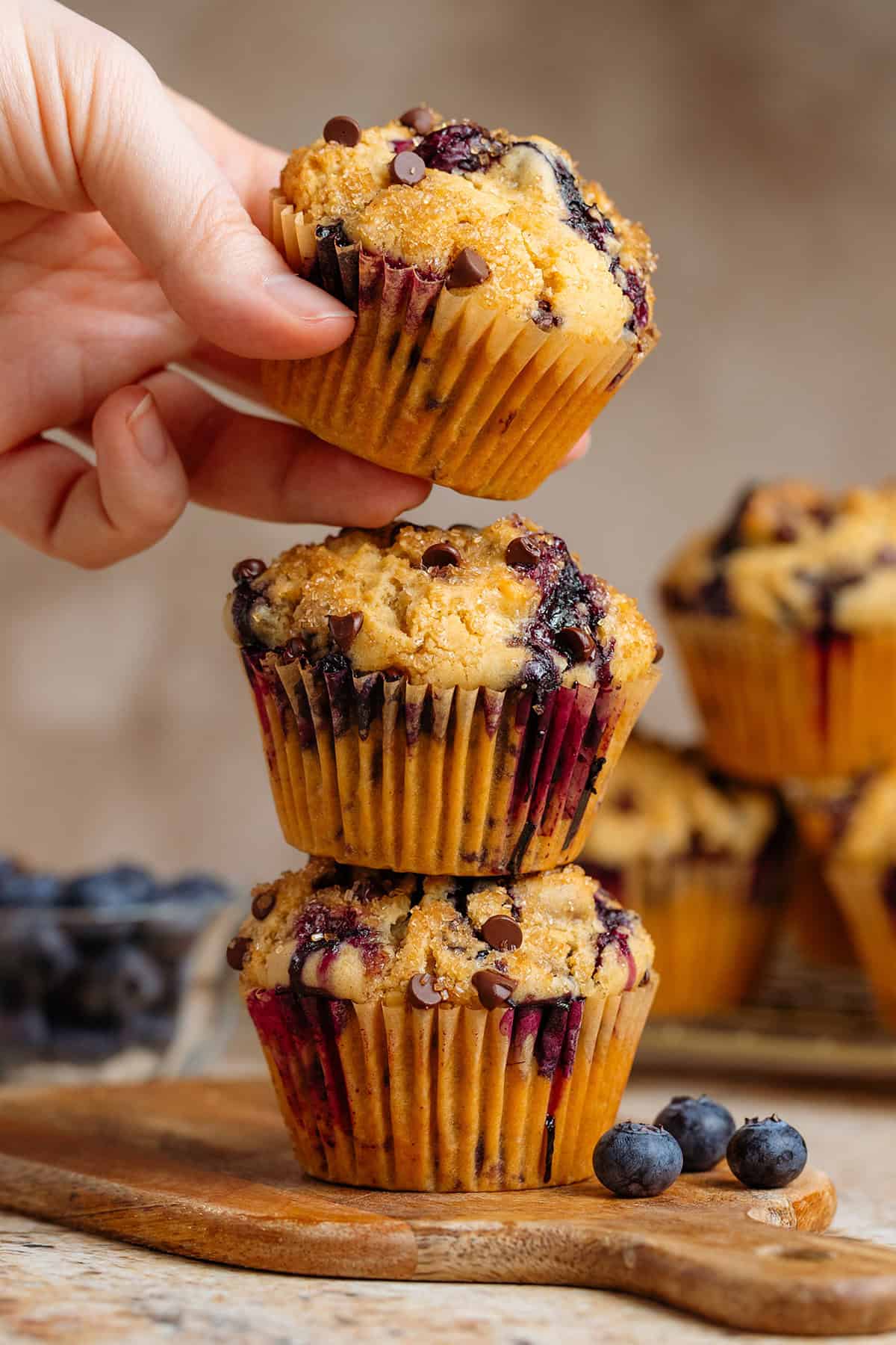 Three blueberry chocolate chip muffins stacked on a small wooden cutting board with a hand grabbing the one on top.