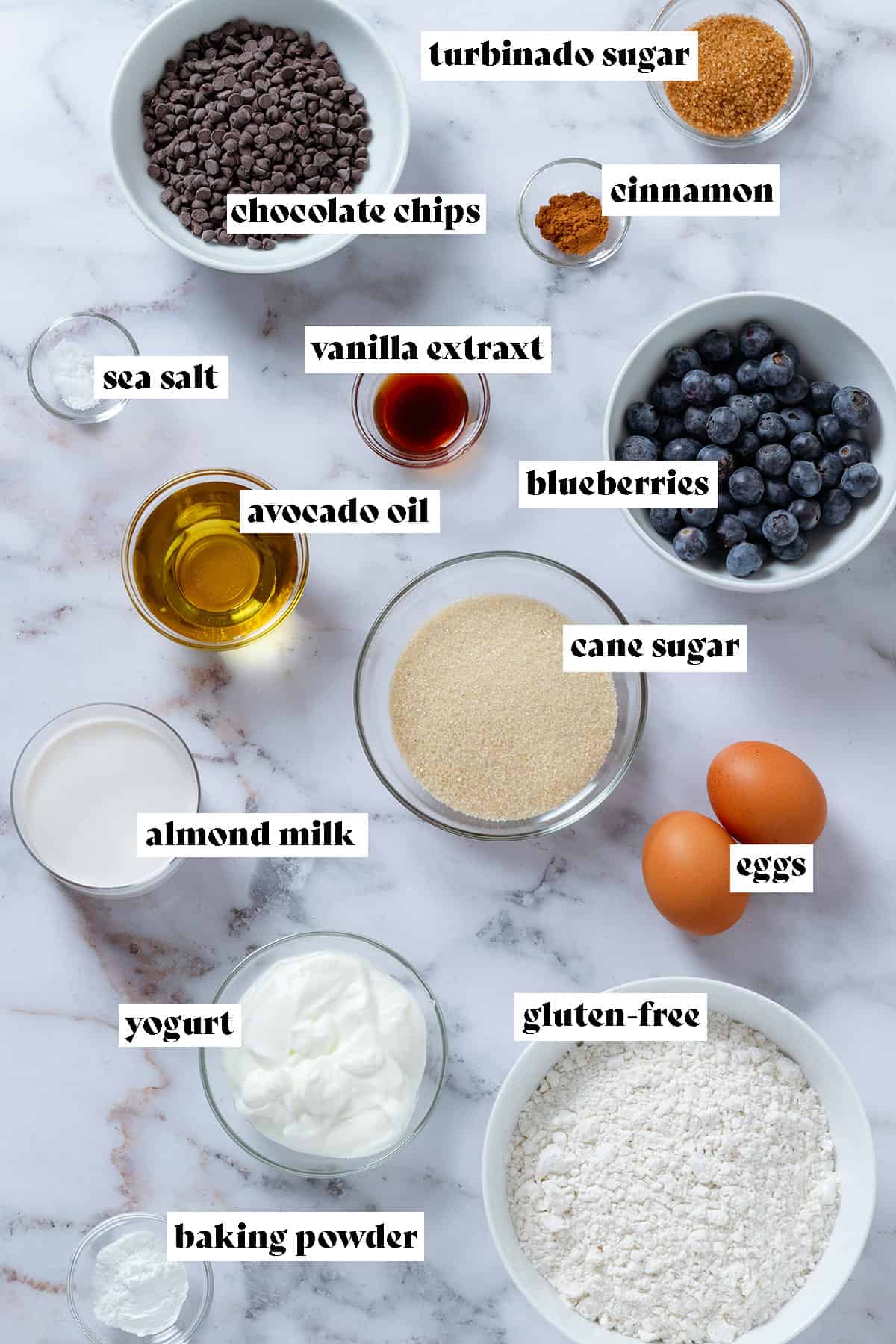 Ingredients like flour, eggs, chocolate chips, blueberries,sugar, and yogurt in glass bowls with text overlay.
