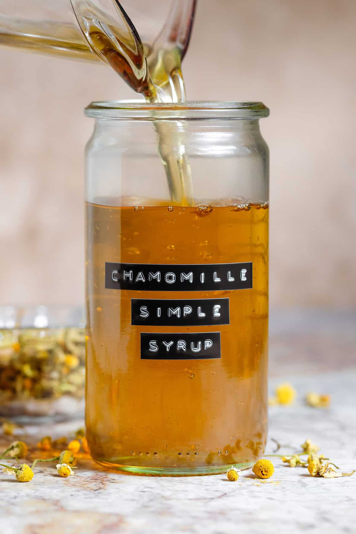 Golden chamomile simple syrup being poured into a tall glass jar with an embossed label.