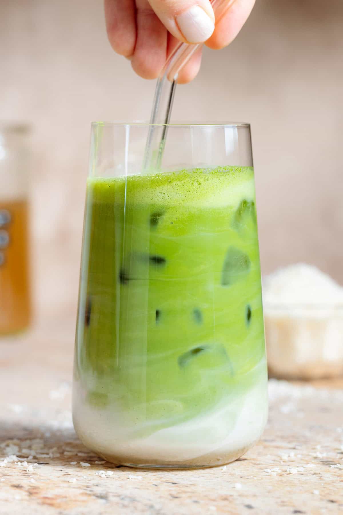 A hand stirring a matcha latte in a tall glass using a glass straw.