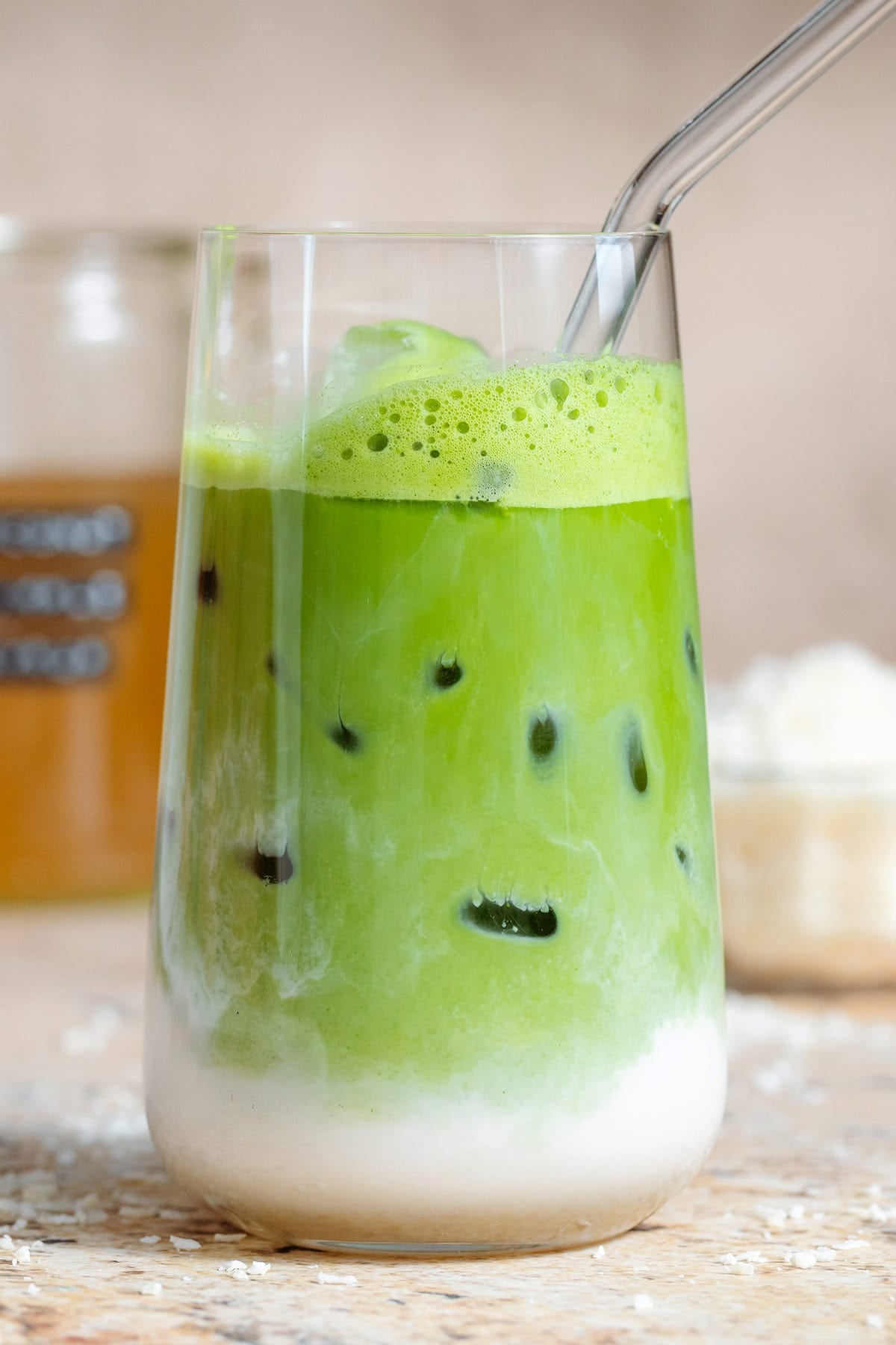 Iced matcha latte in a tall glass with a glass straw and shredded coconut in the background, the milk is clearly separated from the matcha on the bottom half of the glass.
