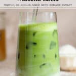 Iced matcha latte in a tall glass with a hand stirring the latte using a glass straw.