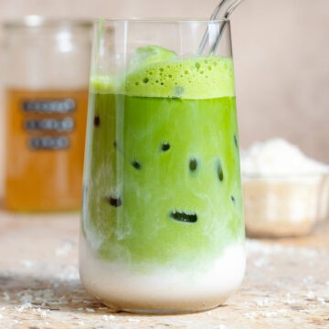 Iced matcha latte in a tall glass with a glass straw and shredded coconut in the background, the milk is clearly separated from the matcha on the bottom half of the glass.