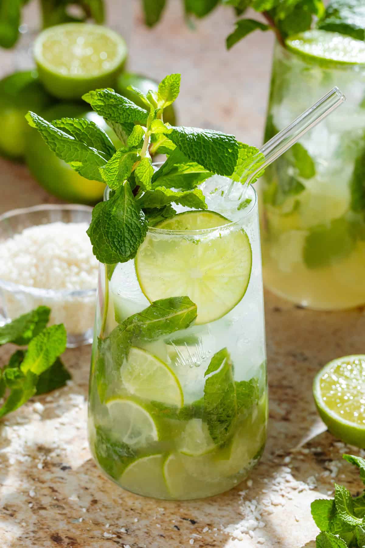 Two tall glasses with coconut mojito garnished with fresh mint leaves, a lime slice, and a glass straw with shredded coconut, limes, and mint in the background.