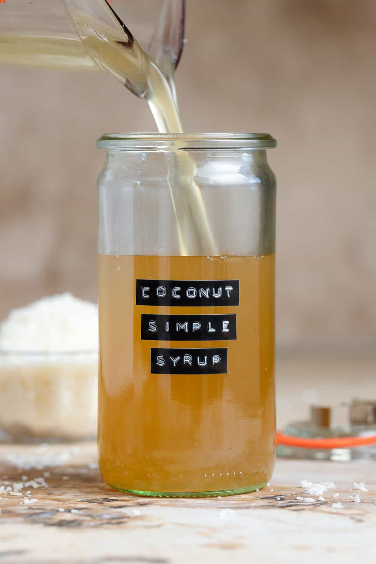 Light yellow coconut simple syrup being poured into a tall glass jar with a black embossed label.