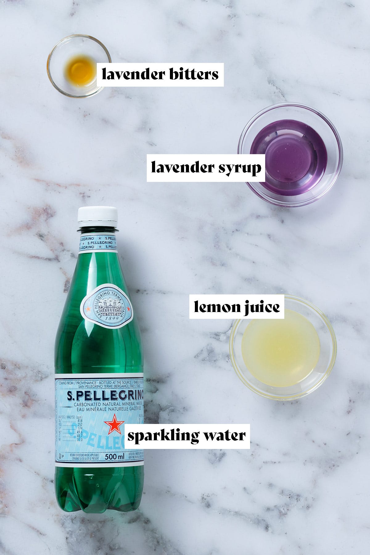 Sparkling water bottle and small glass bowls with lemon juice, lavender syrup, and lavender bitters.
