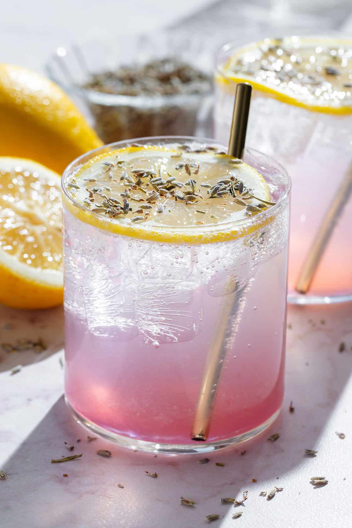 Light pink drinks in short glasses garnished with slices of lemon, dried lavender, and with gold straws.