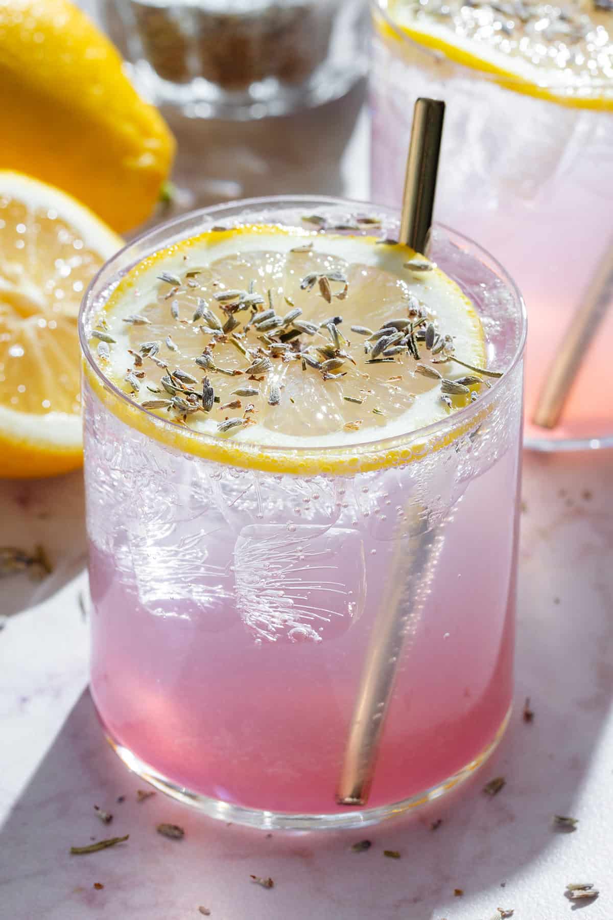 A light pink drink in short glass garnished with a slice of lemon, dried lavender, and with a gold straw.