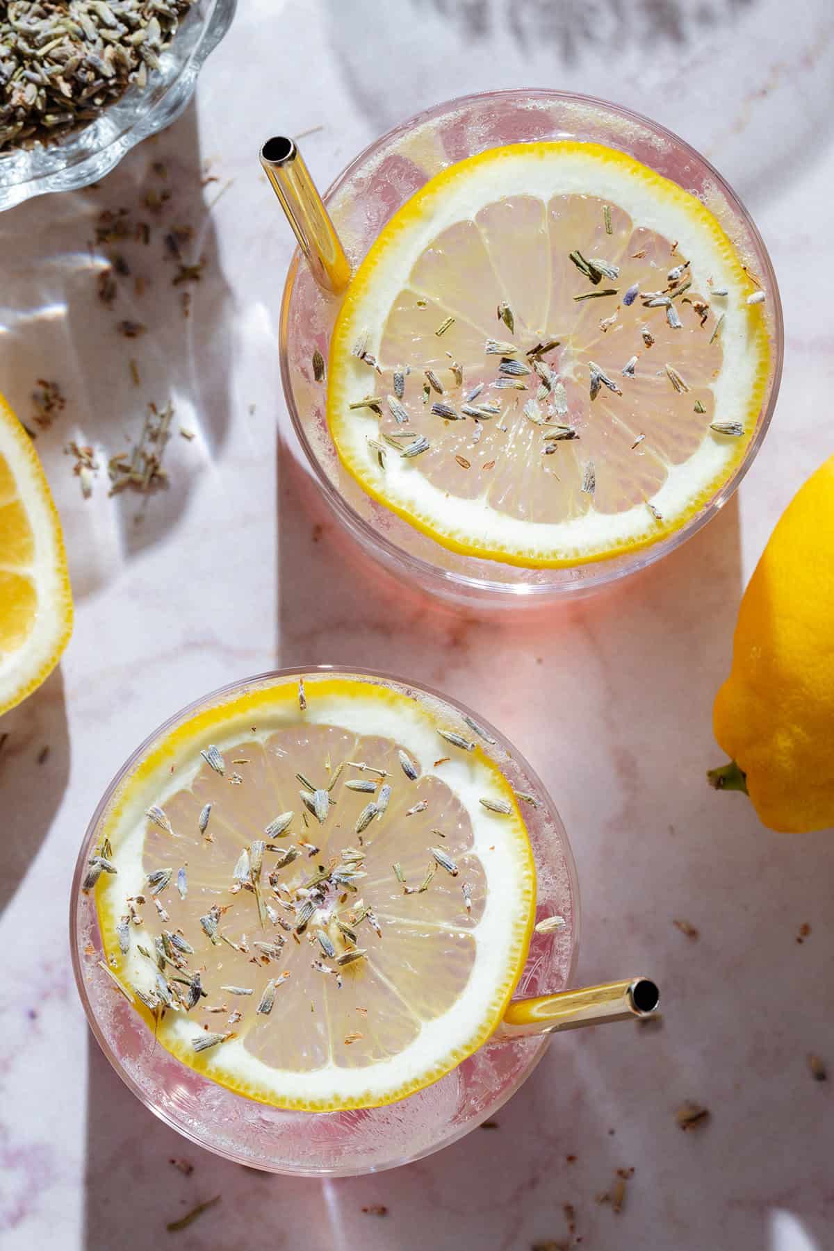 Light pink drinks in short glasses garnished with slices of lemon, dried lavender, and gold straws, with more lemons and lavender around.