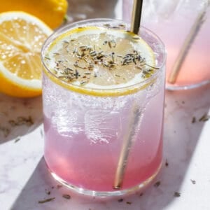 Light pink drinks in short glasses garnished with slices of lemon, dried lavender, and gold straws, with more lemons and lavender around.