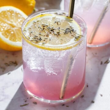 Light pink drinks in short glasses garnished with slices of lemon, dried lavender, and gold straws, with more lemons and lavender around.