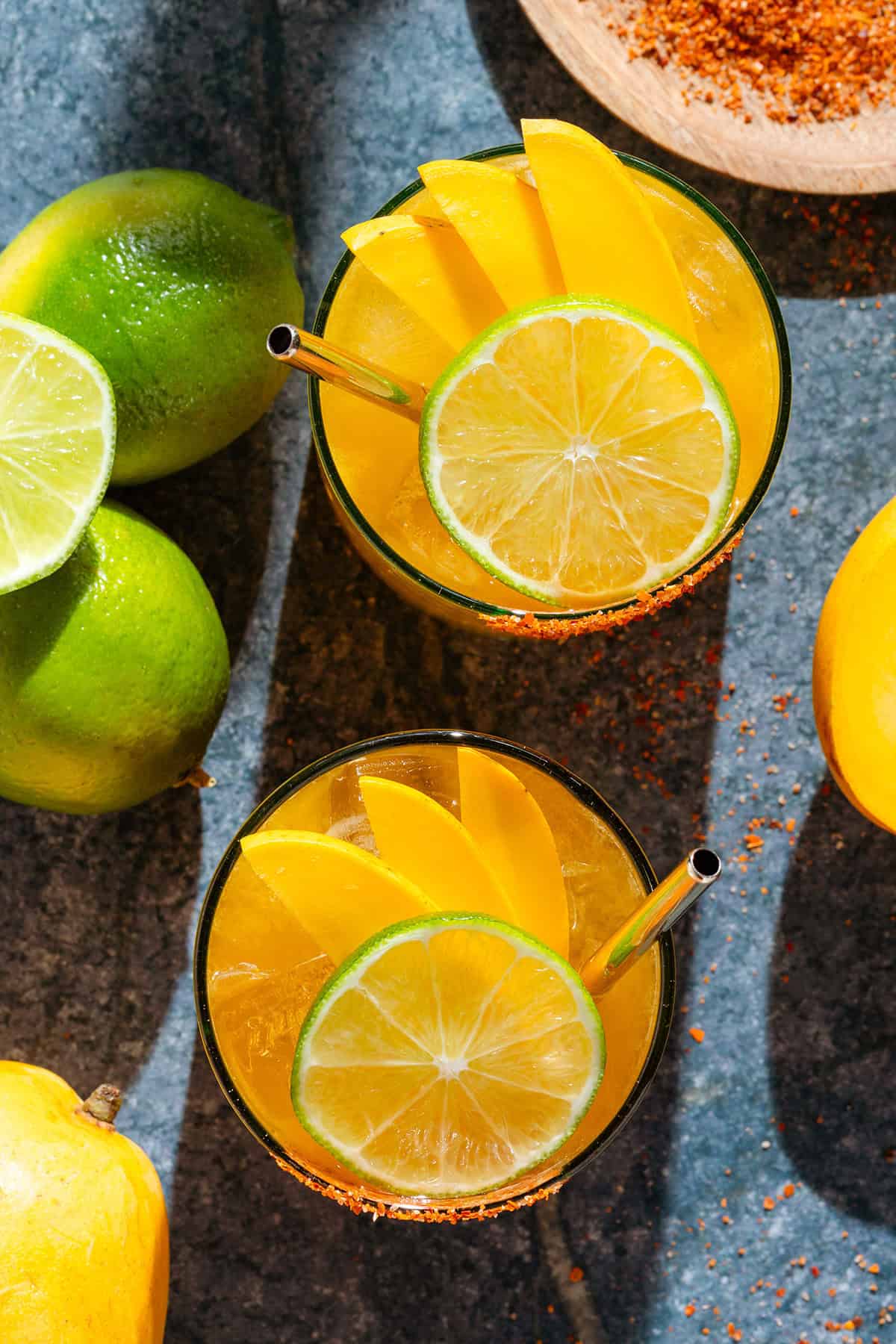 Bright orange mango margarita with ice in two short glasses with tajin rims, garnished with a slices of lime and mango, and with a gold straws with more limes, mangoes, and tajin around.