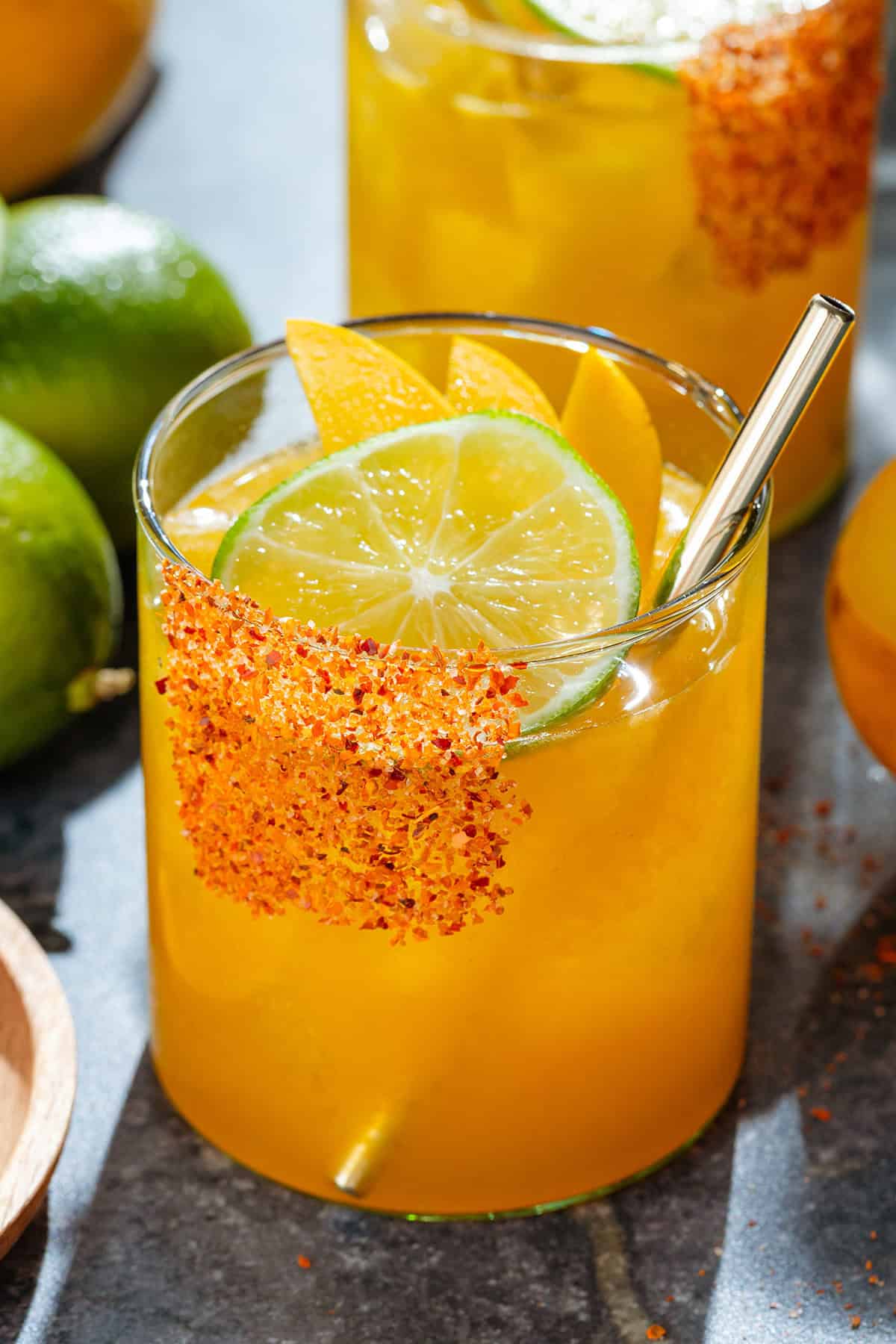 Bright orange mango margarita with ice in a short glass with a tajin rim, garnished with a slices of lime and mango, and with a gold straw.