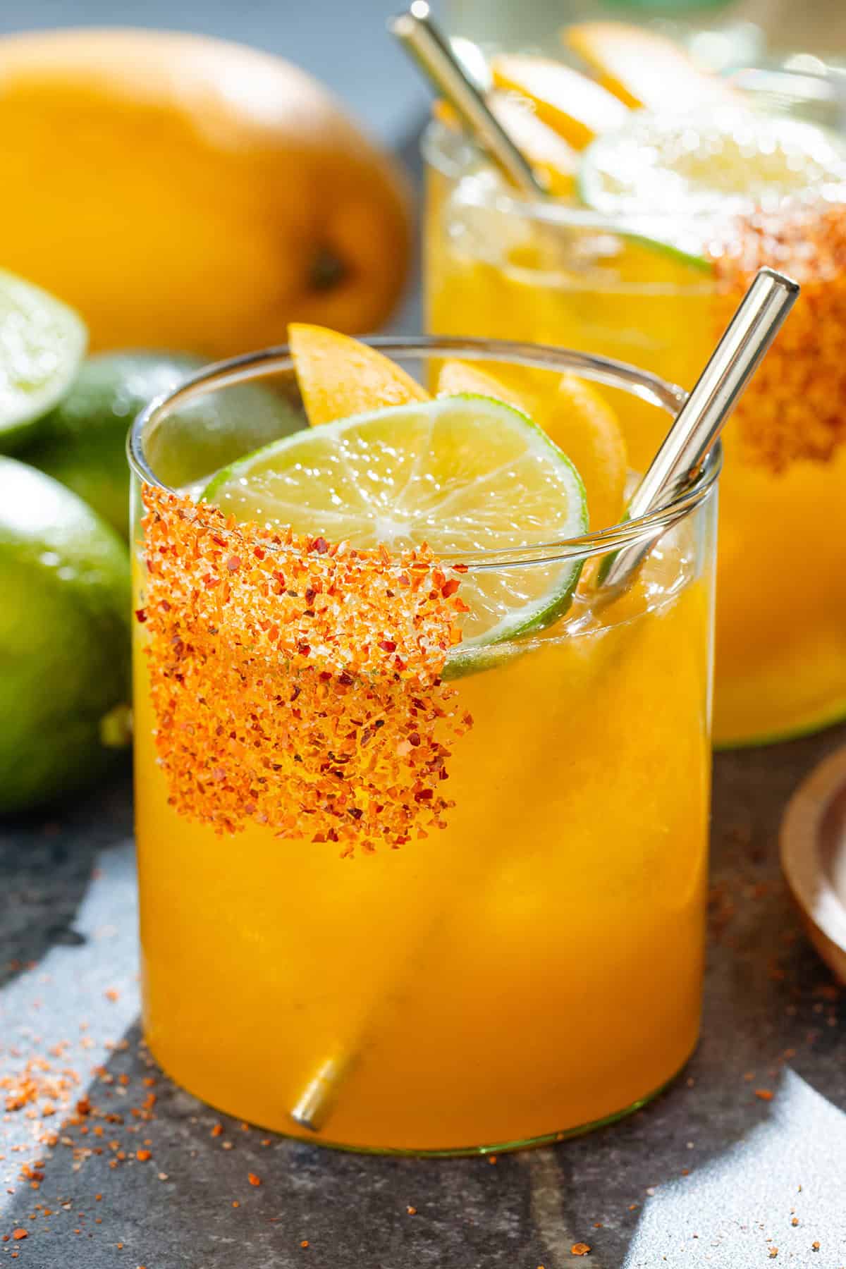 Bright orange mango margarita with ice in a short glass with a tajin rim, garnished with a slices of lime and mango, and with a gold straw.