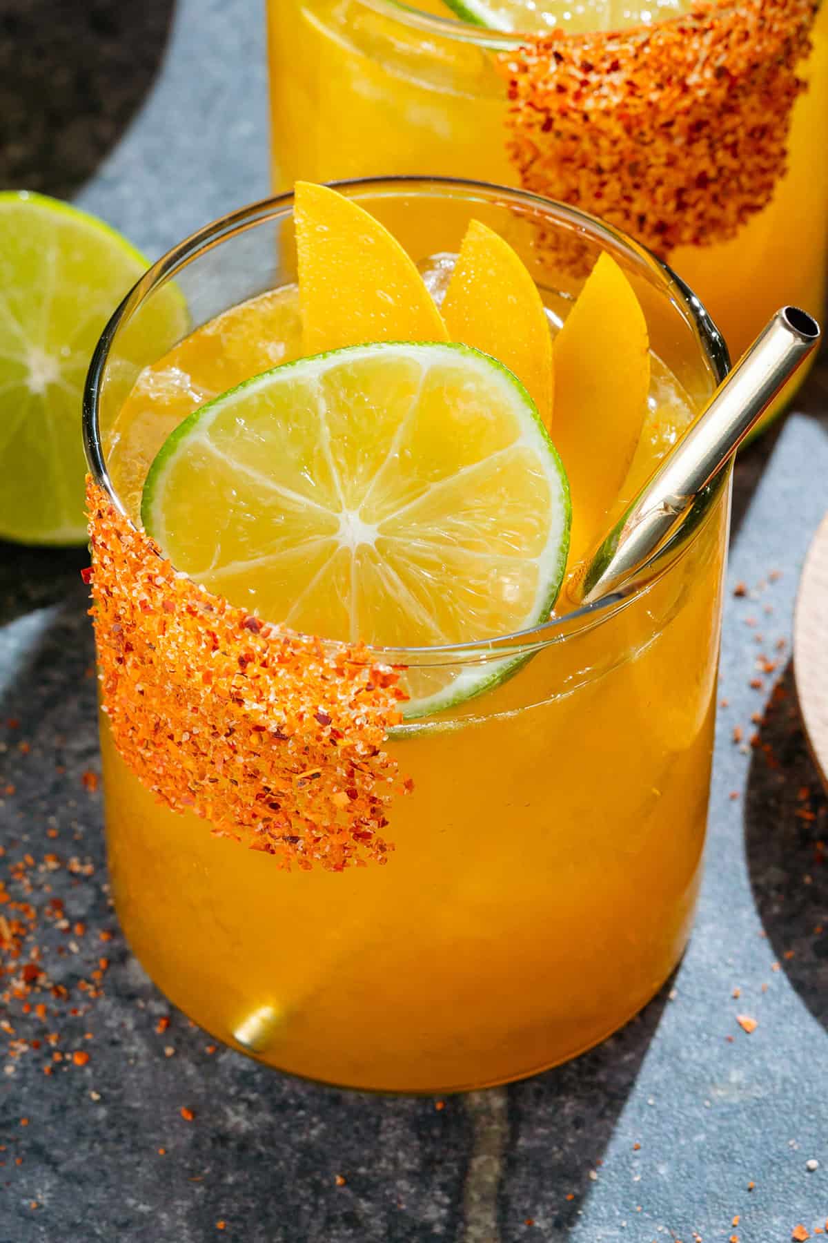 Bright orange mango margarita with ice in a short glass with a tajin rim, garnished with a slices of lime and mango, and with a gold straw.