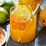 Bright orange mango margarita with ice in a short glass with a tajin rim, garnished with a slices of lime and mango, and with a gold straw.