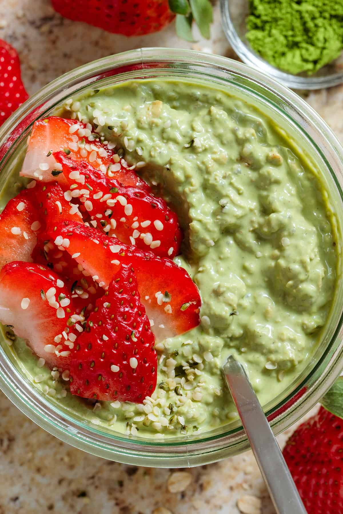Matcha chia pudding in a small glass jar garnished with sliced strawberries and seeds with a spoon in the jar.