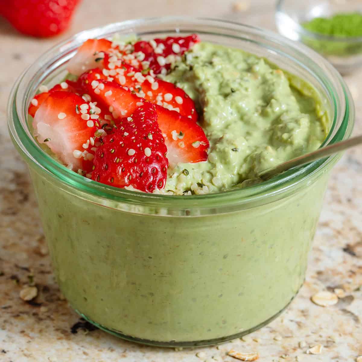 Matcha chia pudding in a small glass jar garnished with sliced strawberries and seeds with a spoon in the jar.