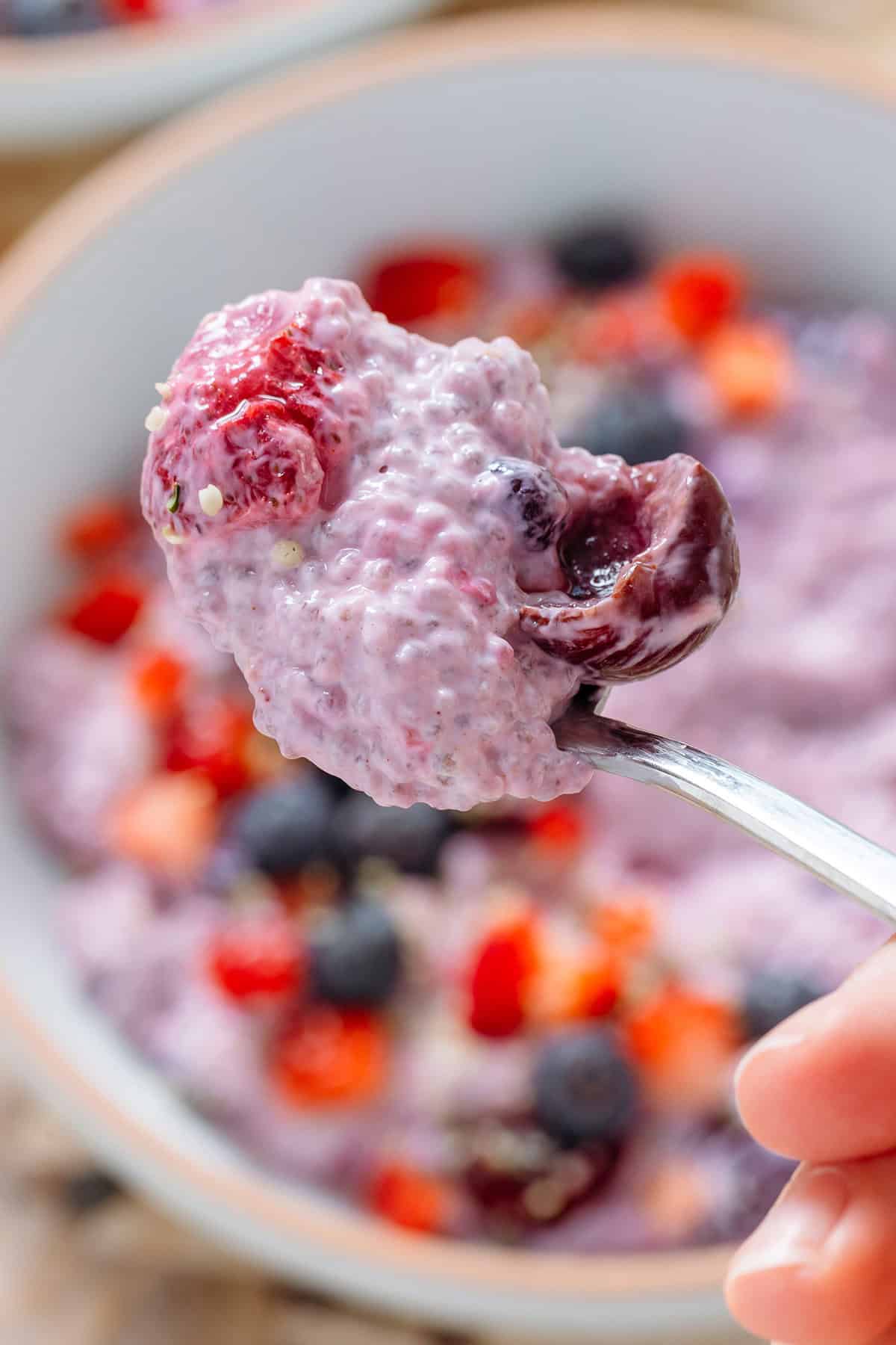 A hand holding a spoon scooping chia pudding with mixed berries over a bowl of the chia pudding.
