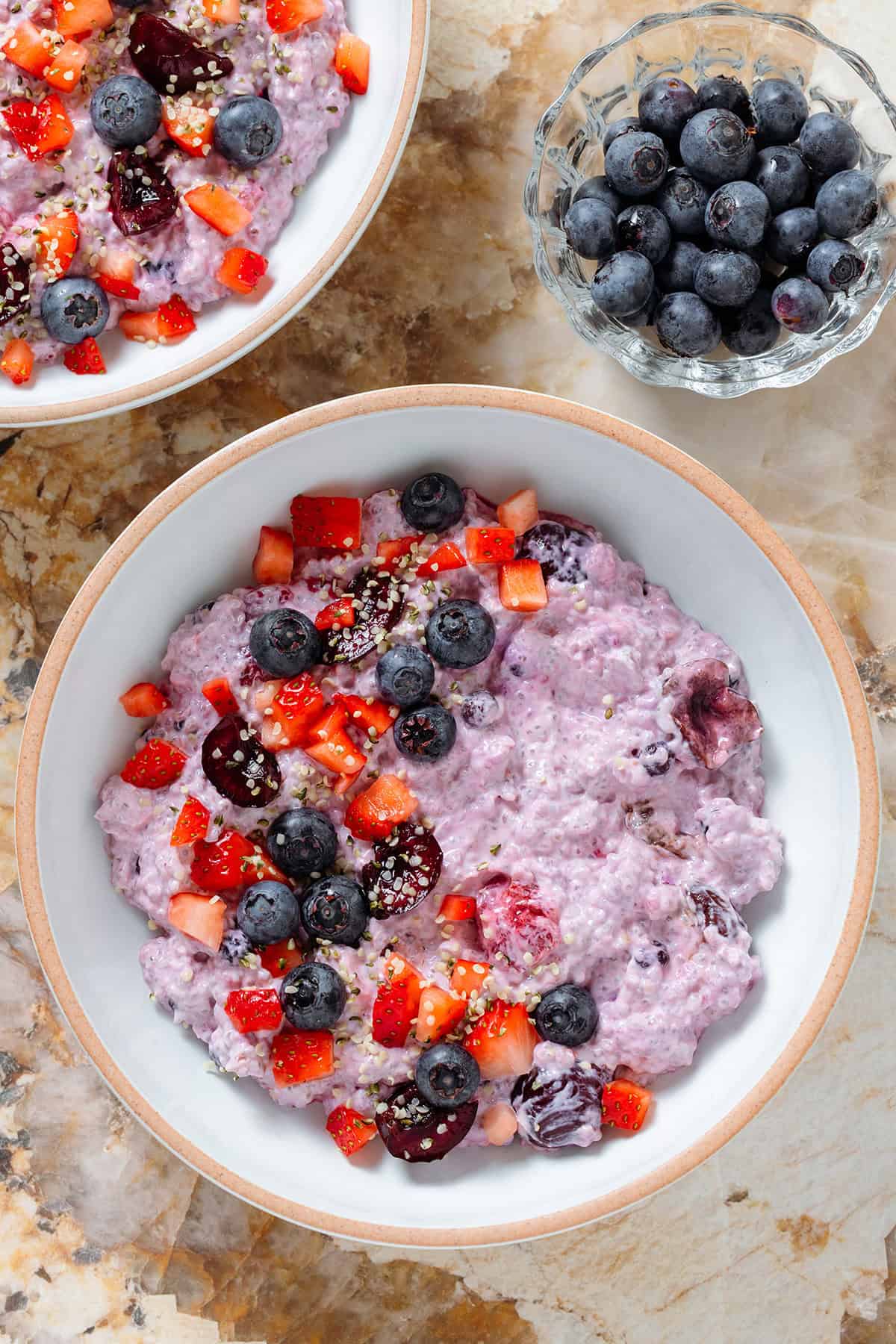 Light purple chia pudding with mixed berries in a low white bowl topped with fresh blueberries, cherries, and chopped strawberries.